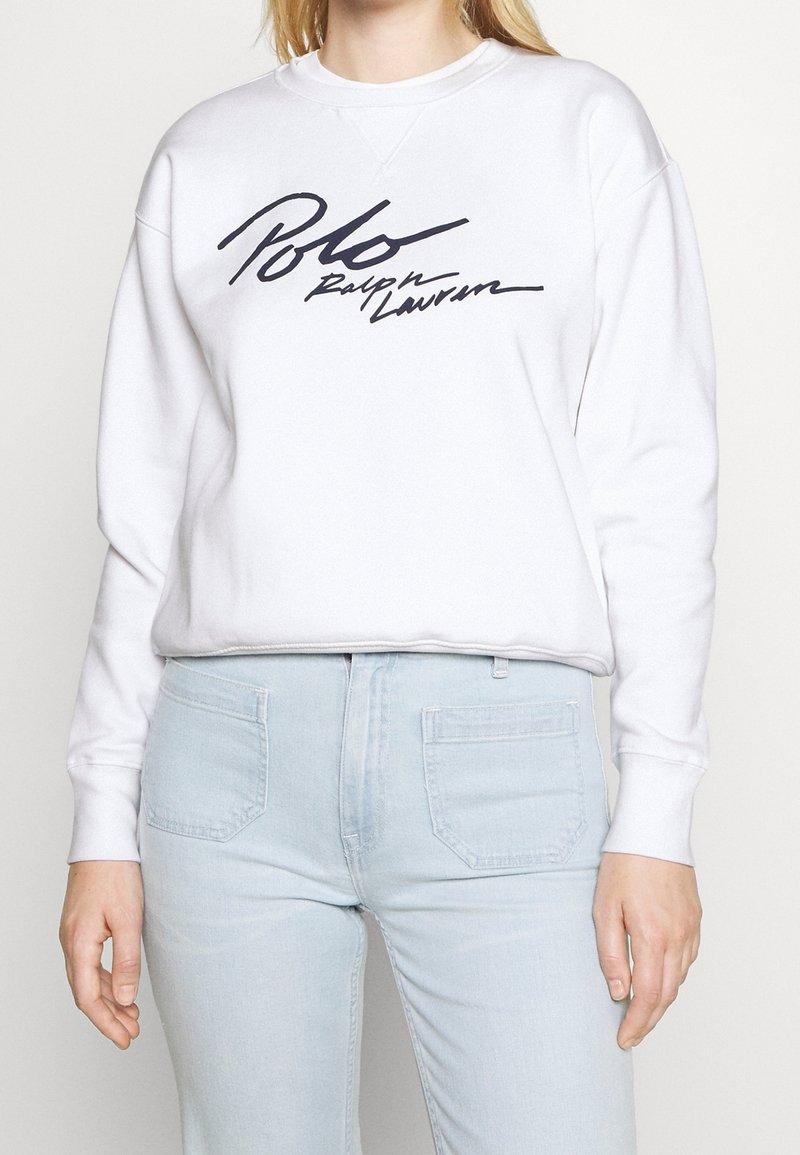 Personne portant un sweat-shirt blanc avec le texte "Polo Ralph Lauren" et un jean bleu clair, debout devant un fond clair uni.