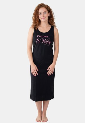 Femme aux cheveux roux bouclés, debout pieds nus, portant une robe noire sans manches avec un texte rose indiquant "Future Wifey" et un graphisme d'anneau.