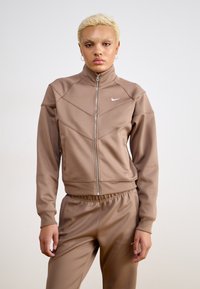 Smeđa jakna na zatvarač s visokim ovratnikom i dizajnom ševrona. Izrađena od glatkog materijala s matching hlačama, s malim logotipom na prsima.