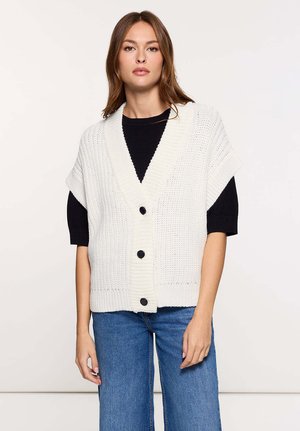 Cárdigan blanco de punto con cuello en V, mangas cortas y tres botones negros en la parte delantera. Combinado con una camiseta negra y pantalones vaqueros.