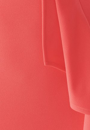 Blusa corallo con una texture liscia e leggera; maniche corte con orlo semplice. Il tessuto appare leggermente trasparente e cade morbido.