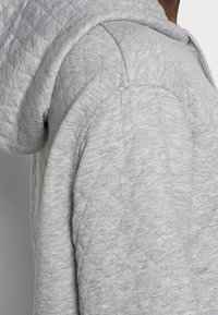 Grå hoodie med quiltad textur och en stor huva. Tyget verkar mjukt och något melerat, med förstärkta axelsömmar.