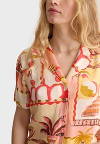 Camisa de manga corta en tejido ligero con un patrón abstracto en tonos rosa y crema que presenta plantas y formas geométricas. Incluye cierre de botones.