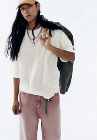 Pullover lejer dintr-un tricotaj subtire, pantaloni roz din denim, jachetă verde cu imprimeu leopard aruncată pe umăr, accente de bijuterii argintii.