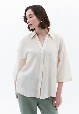 Chemise boutonnée beige clair en tissu texturé, avec un col en V, des manches courtes et une coupe décontractée. Portée avec un pantalon vert.
