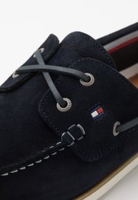 Marineblaue Wildleder-Bootsschuh mit weißen Nähten, Lederschnürsenkeln, Metallösen und einem rot-weiß-blauen Tommy Hilfiger-Logo an der Seite.