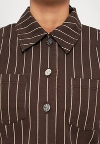 Barna csíkos ing, amely galléros nyakkal, két mellzsebbel és texturált gombokkal rendelkezik. Az anyag vászonra emlékeztető minőségű.