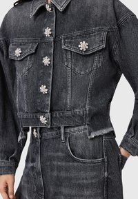 Chaqueta de mezclilla negra cortada con botones de strass en forma de flor, dos bolsillos frontales y dobladillo deshilachado. Textura suave, diseño ajustado.