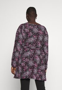 Henkilö, joka käyttää mustaa tunikamekkoa, jossa on vaaleanpunaisia, valkoisia ja violetteja pisteitä, seisoo valkoista taustaa vasten.