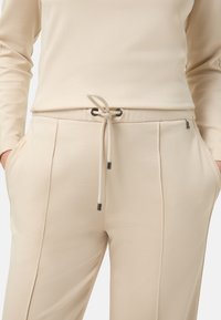 Beżowe spodnie dresowe z sznurkiem w pasie, bocznymi kieszeniami i kontrastującym przeszyciem; gładka tekstura z metalicznymi akcentami na końcach sznurka.