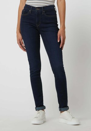 Slim-fit mørkeblå jeans med opsmøgede manchetter og hvide sneakers på en stående person mod en ensfarvet baggrund.