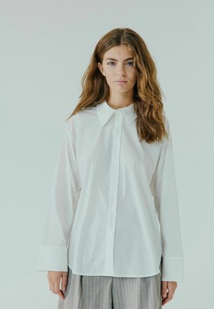 Hvid skjorte med klassisk krave, lange ærmer og en glat tekstur. Kombinationen med grå stribet bukser.