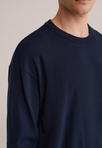 Pull en tricot bleu marine avec un col rond, présentant une coupe décontractée et une texture douce. Couture visible à l'épaule et poignets côtelés.