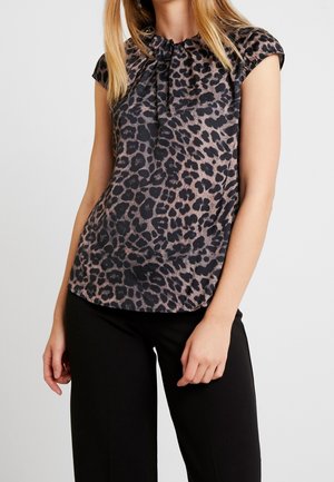 Woman wearing a short sleeve leopard print blouse with gathered neckline and black pants, standing against a plain background.