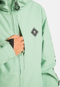 Ręka w zippowanej kieszeni na piersi jasnoszarej kurtki z kapturem i czarną naszywką na piersi.