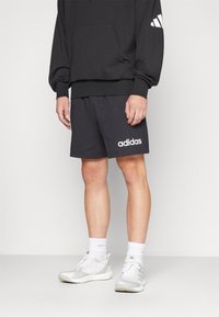 Black hoodie with a front pocket, paired with dark shorts featuring white "adidas" logo. White athletic shoes and socks complete the outfit.