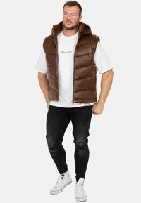 Gilet imbottito marrone con cappuccio, design trapuntato, zip frontale e tasche laterali. Indossato sopra una maglietta bianca e abbinato a jeans neri e sneakers bianche.