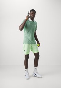 Polo vert avec un col blanc, associé à un short vert néon clair. Le modèle tient une raquette de tennis et une balle, portant des chaussures de sport blanches.