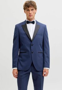 Jauns vīrietis, tērpies pieguļošā zilā smokings ar melniem atloki, baltu kreklu un melnu tauriņu, stāv pret vienkāršu gaišu fonu.