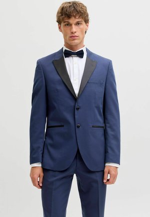 Noor noormees seljas sobiv sinine ülikond mustade kraedega, valgete särgiga ja musta kikilipsuga, seisab ühevärvilise heleda tausta ees.