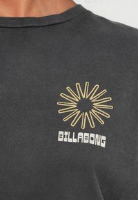 Sötétszürke pamut póló sárga napkitörés grafikával és fehér "BILLABONG" felirattal, kerek nyakkivágással és puha anyaggal.