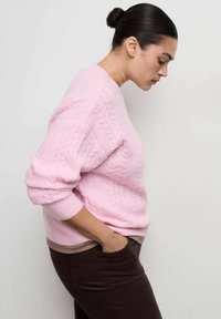 Maglione rosa a trecce con polsini a costine, dal taglio rilassato. Indossato sopra pantaloni marroni, evidenziando la morbida texture e il design.