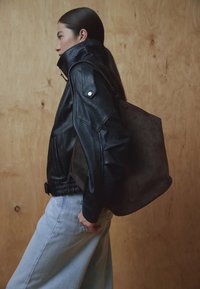 Crna kožna jakna s visokim ovratnikom, s teksturiranim tkanjem i kopčama na drukere. Model nosi tamnocrnu torbu od antilopa i svijetle traperice.