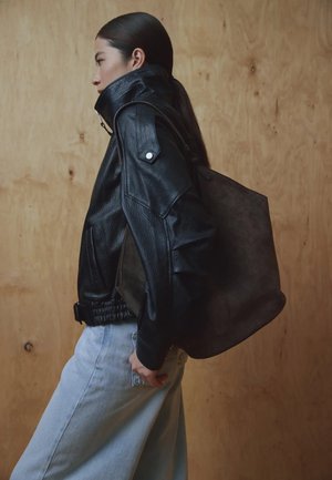 Schwarze Lederjacke mit hohem Kragen, aus strukturiertem Stoff mit Druckknöpfen. Das Modell trägt eine dunkelbraune Wildleder-Tasche und helle Jeans aus Denim.