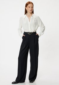 Weiße Bluse mit langen Ärmeln und Kragen, kombiniert mit hochtaillierten schwarzen weiten Hosen, die einen schwarzen Gürtel mit einer runden Schnalle haben.