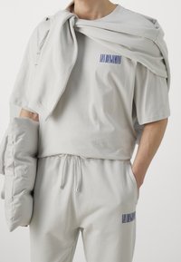 Svetle šedý leisurewear set, který zahrnuje top s krátkým rukávem a kalhoty na elastický pás, oboje s tištěným textem a měkkou, hladkou texturou.