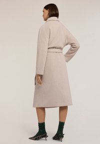 Gabardina de lana beige con cintura ceñida, mangas largas y dobladillo ligeramente acampanado. Combinada con calcetas verdes de canalé y zapatos de tacón negros.