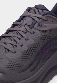 Κοντινή λήψη ενός γκρι παπουτσιού τρεξίματος Hoka με γκρι κορδόνια, υφασμάτινο πλέγμα και μοβ λεπτομέρειες λογότυπου στο πλάι και στη γλώσσα.