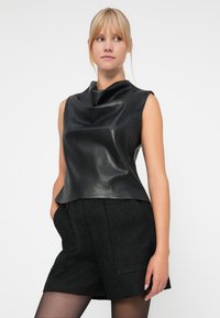 Top nero senza maniche realizzato in materiale liscio e lucido con scollatura drappeggiata; abbinato a pantaloni corti neri con texture.