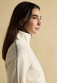 Beżowy sweter na zamek z wysokim kołnierzem, z ciemnoniebieskim paskiem po bokach i gładką teksturą, pokazany z boku na neutralnym tle.