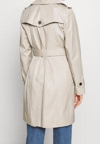Beżowy płaszcz trencz z paskiem w talii, rozcięciem z tyłu i naramiennikami. Posiada duże guziki oraz gładką, wodoodporną fakturę.