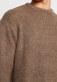 Närbild av en person som bär en tjock, brun, ribbad stickad tröja med rund halsringning.