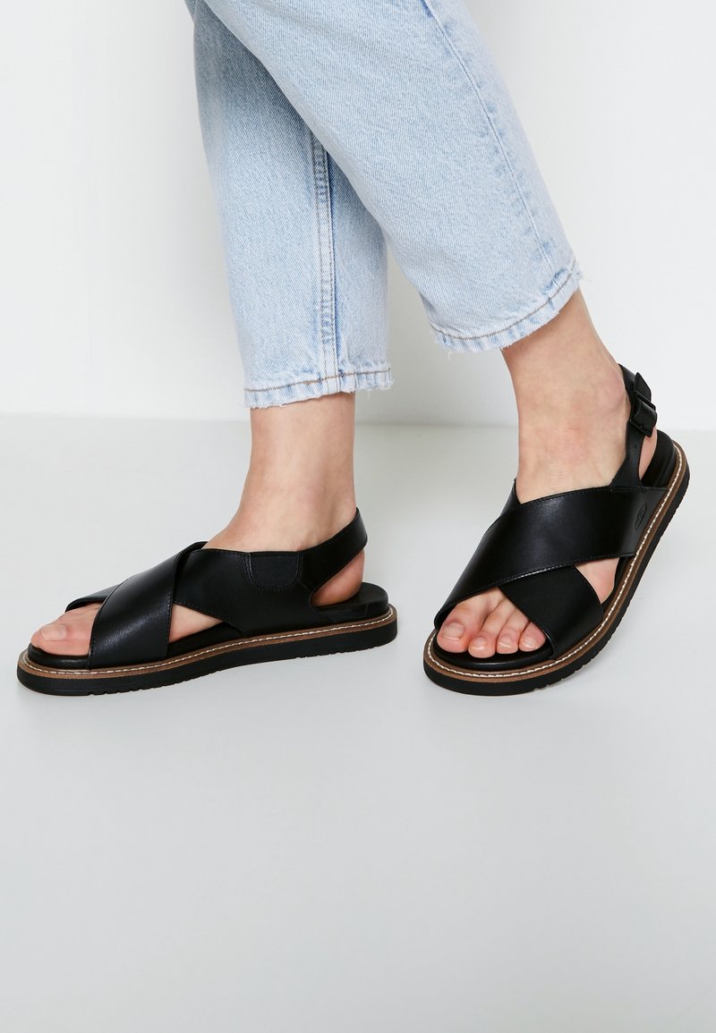 Pieds portant des sandales en cuir noir à brides croisées avec boucle à la cheville, associées à un jean court bleu clair, debout sur une surface blanche.