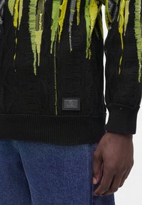 Gros plan d'un pull texturé noir avec des motifs verticaux verts et jaunes porté avec un jean bleu, montrant le bas du torse et la main droite.