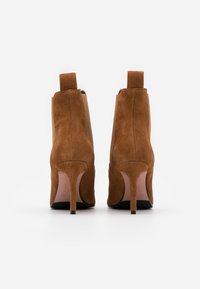 Pari saapikkaita ruskeasta mokasta, korkokengät nilkkasaappaissa, katsottuna takaa valkoista taustaa vasten.