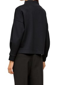 Sudadera corta negra con cuello alto, hombros caídos y puños de canalé. Textura de tela suave, diseño minimalista, sin logotipos visibles.