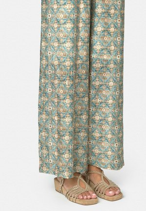 Pieds portant des sandales ouvertes beige ornées de strass, partiellement couverts par un pantalon long bleu sarcelle à motif floral complexe.