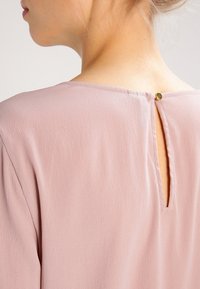 Donna che indossa un top testurizzato di un rosa chiaro con un piccolo'apertura a chiave fissata da un singolo bottone dorato nella parte posteriore del collo.