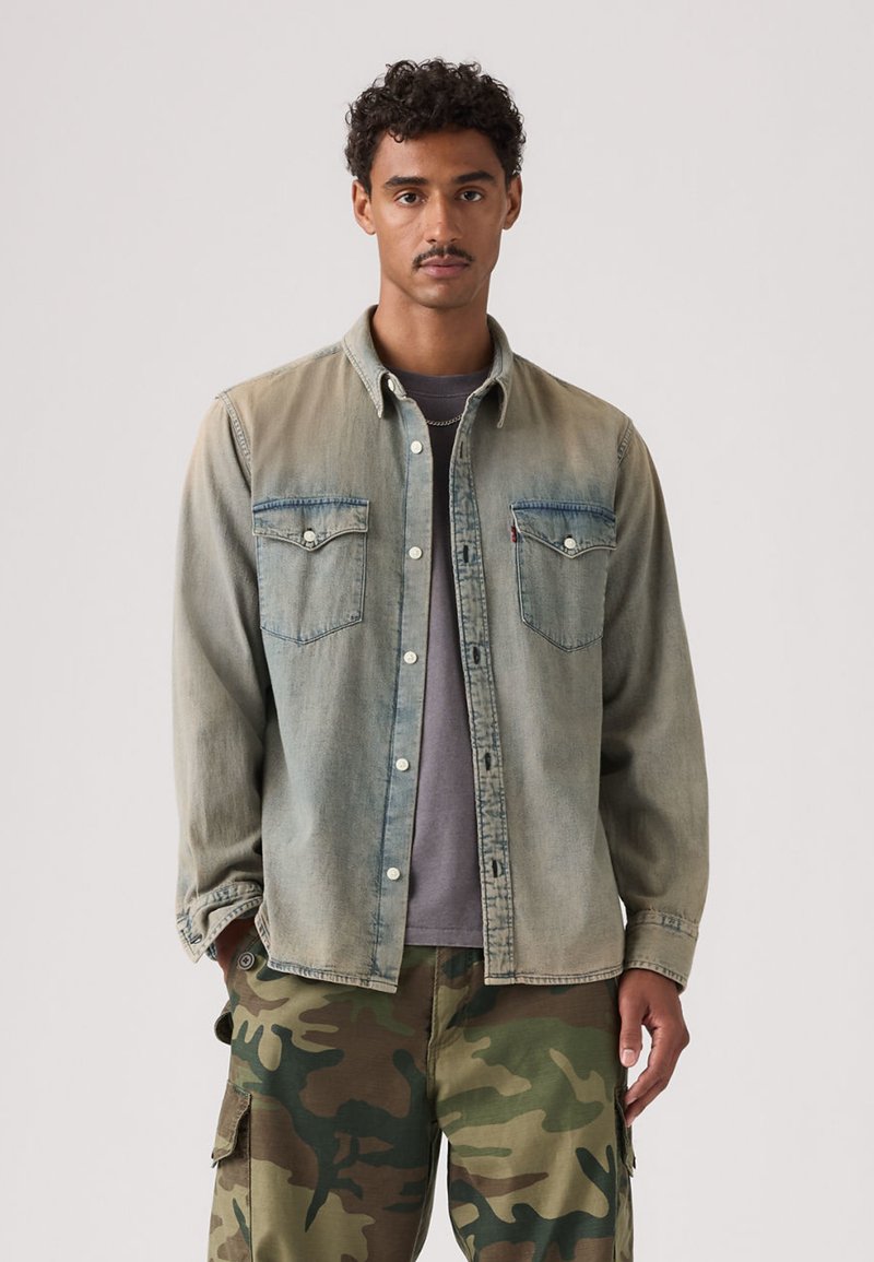 Νεαρός άνδρας με σγουρά μαλλιά, ντυμένος με ξεθωριασμένο τζιν πουκάμισο πάνω από γκρι μπλουζάκι και καμουφλαρισμένο παντελόνι cargo, στέκεται μπροστά σε ένα απλό φόντο.