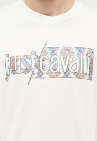 Λευκό μπλουζάκι με το λογότυπο "Just Cavalli" που περιλαμβάνει σχέδια με λουλούδια και ανανά μέσα σε γράμματα με περίγραμμα στο στήθος.