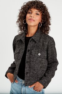 Giacca cropped a trama bianco e nero con chiusura a bottoni, due tasche sul petto e colletto classico, abbinata a una maglietta nera e jeans blu.