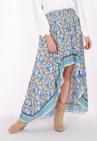 Blumen-Maxi-Rock in Pastellfarben mit blauen und gelben Akzenten, mit einem hoch-niedrig geschnittenen Saum und texturiertem Stoff. Kombiniert mit braunen Stiefeletten.