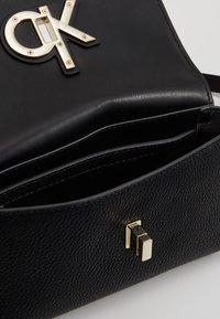 Černá kabelka z texturované kůže s výrazným zlatým logem "CK" a kovovým klipem, uvnitř s otevřenou přihrádkou.
