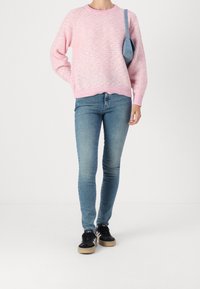 Kobieta ubrana w różowy sweter z dzianiny, niebieskie slim fit dżinsy, czarne trampki z białymi paskami i nosząca małą niebieską torbę na ramię.