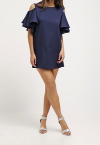 Robe bleu marine à épaules dénudées avec des manches à volants superposés, fabriquée en un matériau lisse. Associée à des sandales à talons hauts en argent métallique.