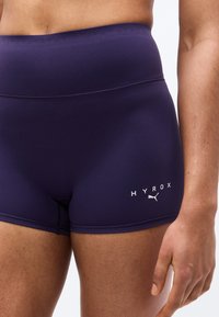 Tuvplāns cilvēkam, kurš valkā tumši zilas sporta šortus ar augstu jostasvietu un baltu "HYROX" un Puma logo uz kreisās augšstilba puses.